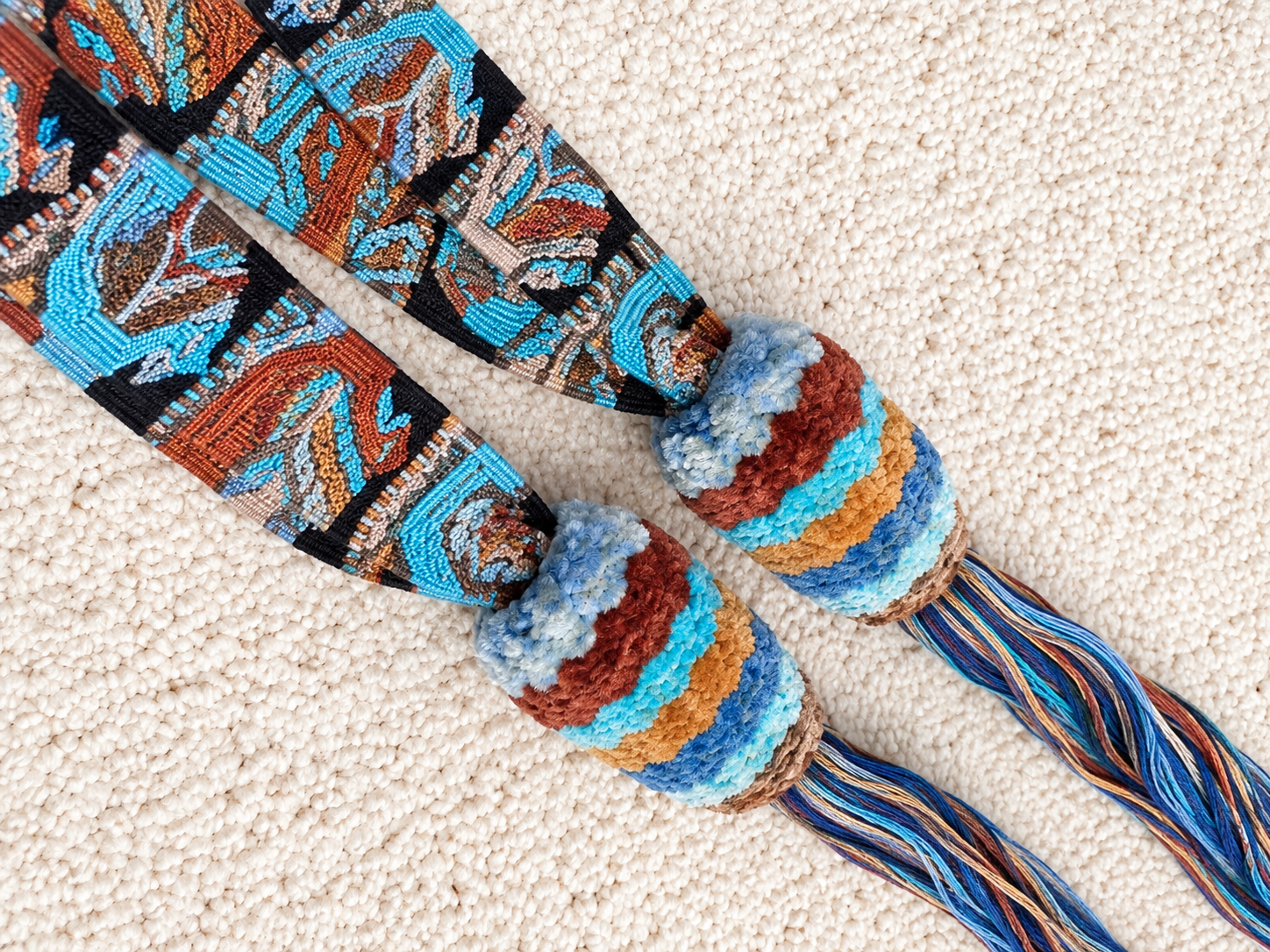 A pair of colorful woven rope accessories with striped knotted loops and patterned hanging strands, laid flat on a textured cream carpet.