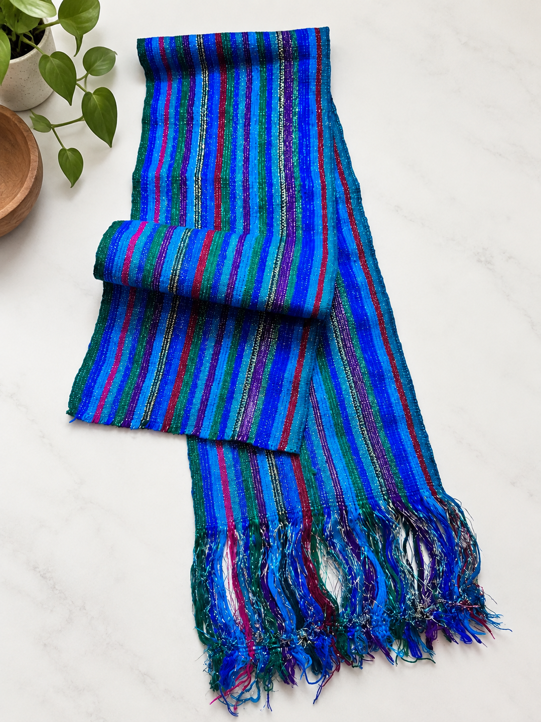 A multicolor striped woven scarf with fringed ends, displayed on a light marble surface next to a small potted plant.