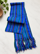 A multicolor striped woven scarf with fringed ends, displayed on a light marble surface next to a small potted plant.