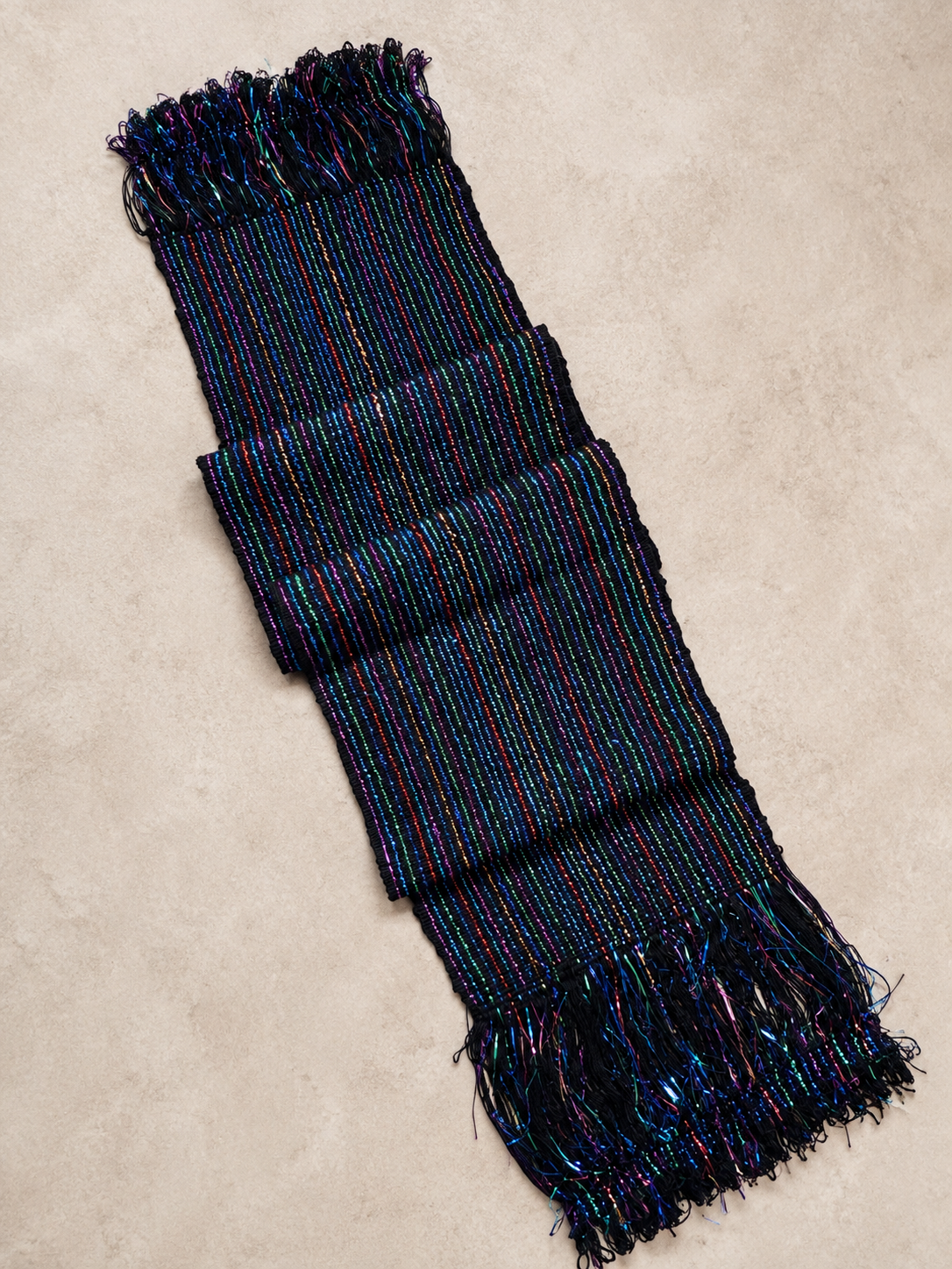 A dark woven scarf with shimmering multicolor threads and fringed ends, laid flat on a textured beige surface.