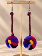A pair of colorful pom-pom dangle earrings hanging from a gold display stand. The earrings feature round, fuzzy pom-poms in a multicolor pattern of purple, pink, blue, yellow, and black, suspended from dark purple braided cords with silver fishhook ear wires.