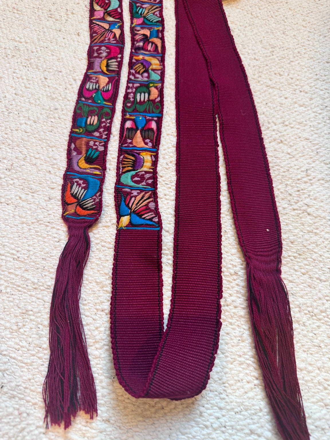 A long burgundy ribbon or strap laid flat on a textured cream surface, with two narrow embroidered vertical panels near the top showing multicolored floral and geometric patterns. The ribbon ends are fringed and the main body is solid burgundy.