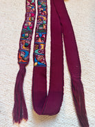 A long burgundy ribbon or strap laid flat on a textured cream surface, with two narrow embroidered vertical panels near the top showing multicolored floral and geometric patterns. The ribbon ends are fringed and the main body is solid burgundy.